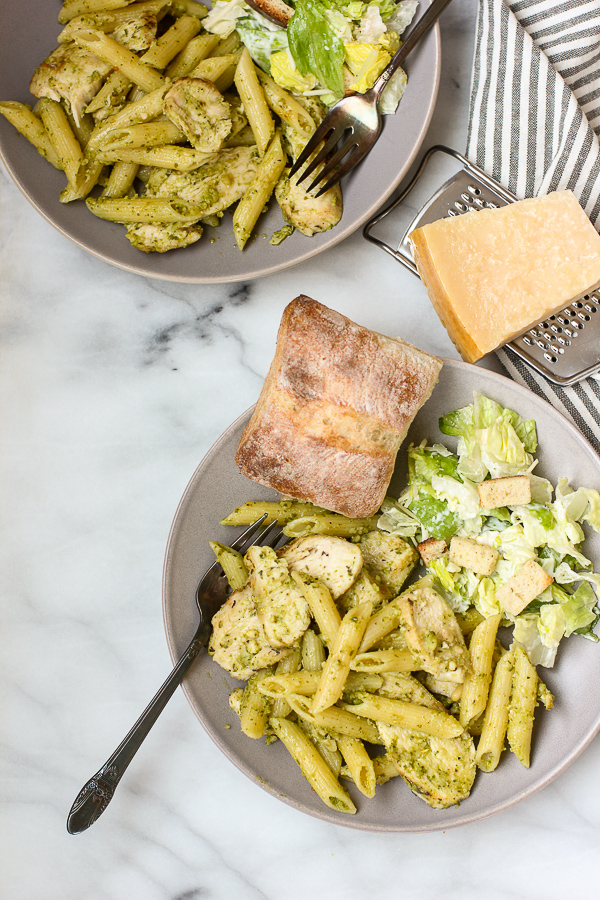 A close-up view of two plates of pesto pasta with chicken, a side salad, and a loaf of bread on a marble surface.
